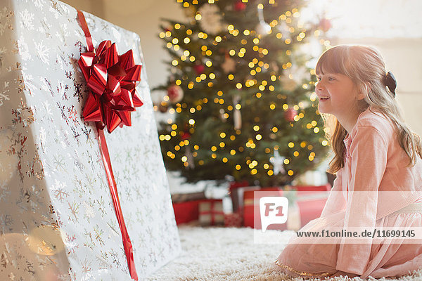 Mädchen lächelt in Erwartung eines großen Weihnachtsgeschenks am Weihnachtsbaum