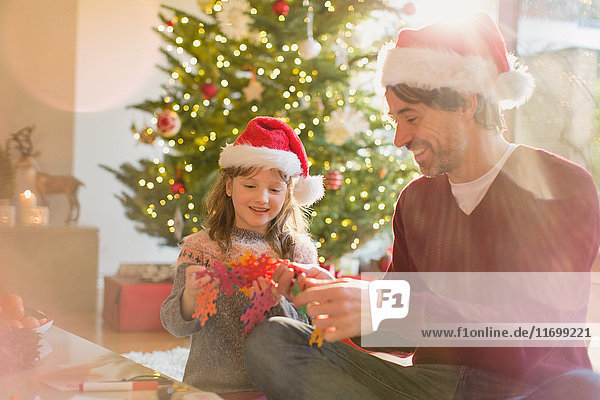 Father and daughter wearing Santa hats and holding paper snowflakes near Christmas tree