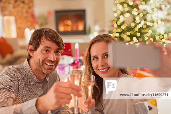 Lächelndes Paar stößt mit Sektflöten an und macht ein Selfie am Weihnachtstisch