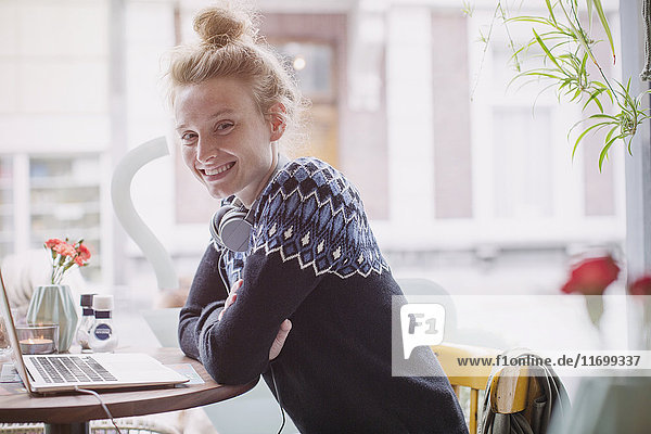 Porträt einer lächelnden jungen Frau mit Kopfhörern am Laptop im Caféfenster