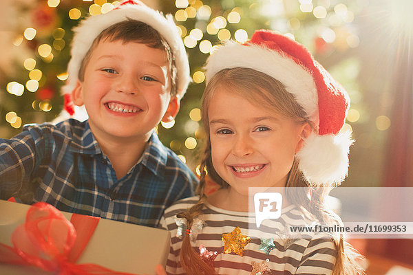 Porträt eines lächelnden  begeisterten Geschwisterpaares mit Weihnachtsmannmützen und Weihnachtsgeschenk