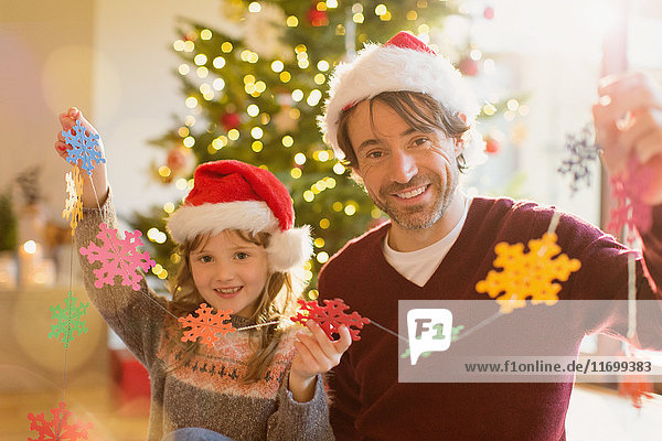 Porträt eines lächelnden Vaters und einer lächelnden Tochter mit Weihnachtsmannmützen  die eine Kette von Papierschneeflocken halten