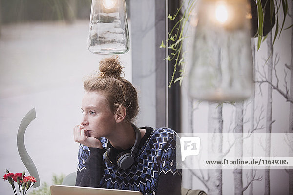 Nachdenkliche junge Frau mit Kopfhörern am Laptop  die aus dem Fenster eines Cafés schaut