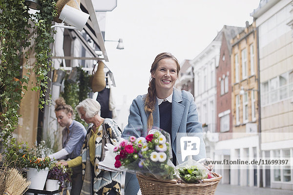 Smiling woman riding bicycle with flowers in basket on city street