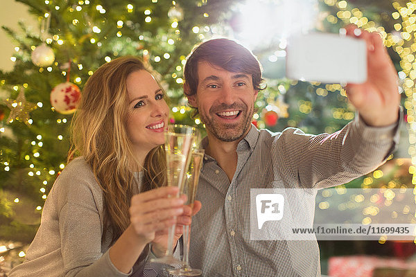Lächelndes Paar stößt mit Sektflöten an und macht ein Selfie vor dem Weihnachtsbaum