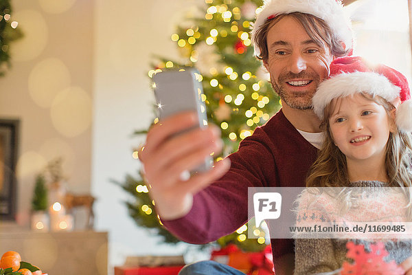 Lächelnder Vater und Tochter mit Weihnachtsmannmützen machen ein Selfie mit einem Fotohandy