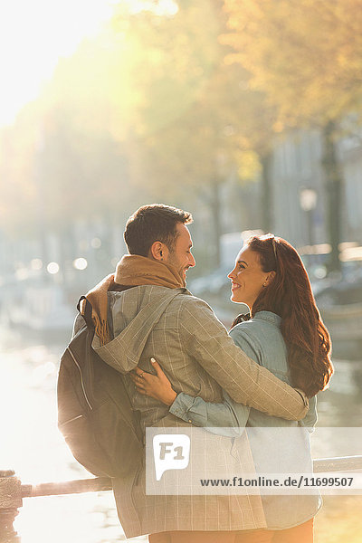 Lächelndes junges Paar  das sich auf der sonnigen Herbstbrücke über den Kanal umarmt.