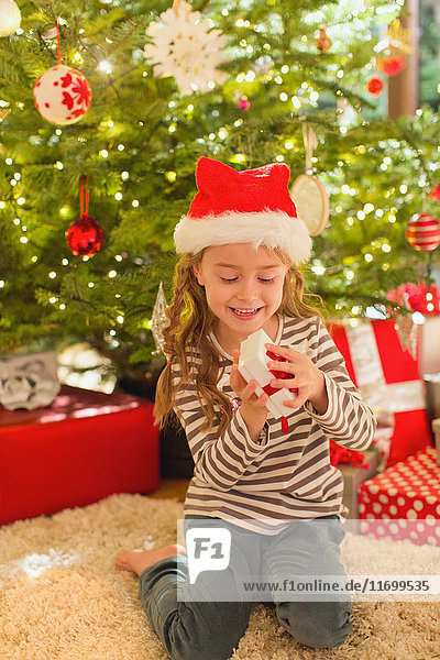 Lächelndes Mädchen mit Weihnachtsmannmütze öffnet ein Geschenk vor dem Weihnachtsbaum