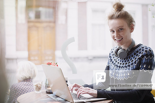 Porträt einer lächelnden jungen Frau  die in einem Caféfenster einen Laptop benutzt