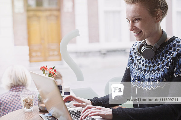 Lächelnde junge Frau mit Kopfhörern und Laptop im Fenster eines Straßencafés
