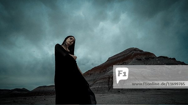 Moody Porträt der Frau in langen schwarzen Kleid in der Nähe von großen Rock-Formation mit grauen Wolken bewegen Overhead