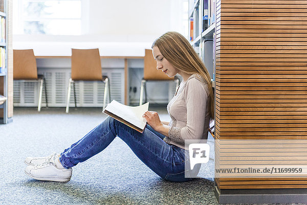 Teenagermädchen sitzt auf dem Boden in einer öffentlichen Bibliothek und liest Bücher.