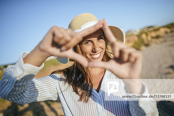 Porträt einer lächelnden jungen Frau  die am Strand einen Fingerrahmen anfertigt.