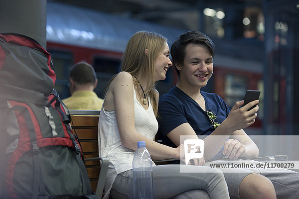 Young couple with backpacks sitting at rail station using smartphone