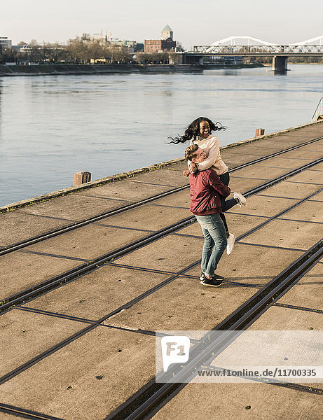Junger Mann hebt junge lachende Frau am Flussufer an