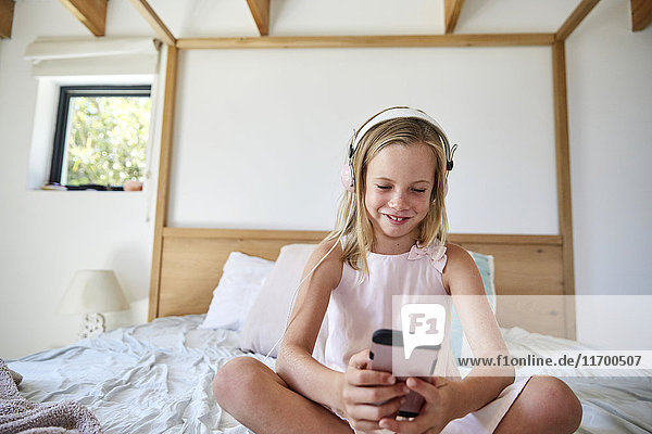 Lächelndes kleines Mädchen sitzt zu Hause auf dem Bett und hört Musik mit Kopfhörern.