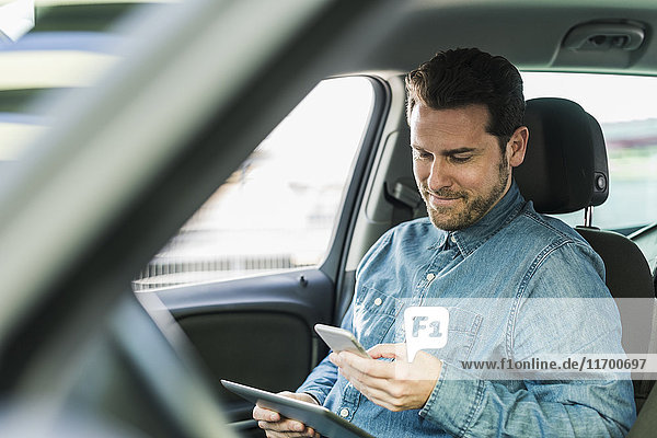 Geschäftsmann im Auto sitzend mit Smartphone und digitalem Tablett