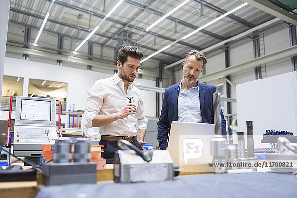Two men at table in factory shop floor looking at laptop