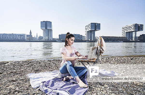 Germany  Cologne  young woman using laptop at River Rhine