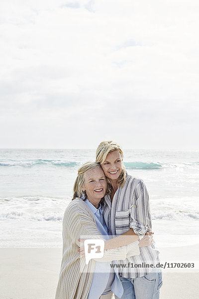 Seniorin und ihre erwachsene Tochter stehen am Strand und umarmen sich.