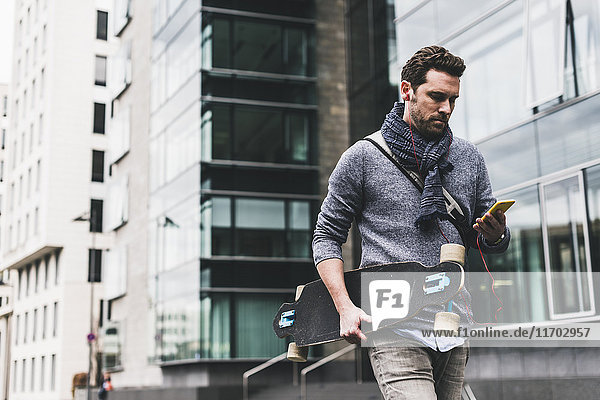 Businessman carrying skateboard  using smartphone and earphones