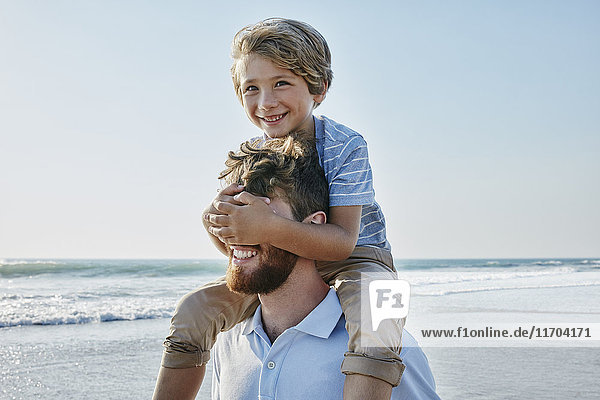 Father carrying son piggyback on the beach  son covering father's eyes