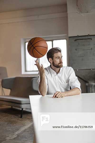 Porträt eines nachdenklichen jungen Geschäftsmannes  der am Tisch in einem Loft sitzt und Basketball auf seinem Finger balanciert.