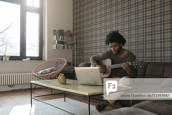 Man sitting in living room on sofa playing guitar in front of laptop
