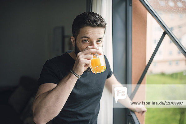 Junger Mann trinkt einen Orangensaft am Fenster.