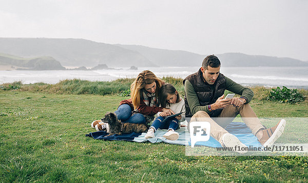 Familie mit Hund  der an der Küste auf einer Decke sitzt und drahtlose Geräte benutzt.