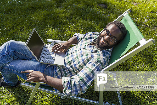 Smiling man with laptop sitting on deck chair on a meadow looking up to camera