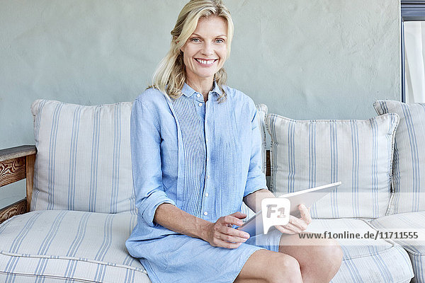 Porträt einer lächelnden blonden Frau auf der Couch mit Tablette