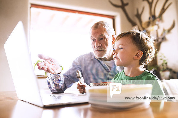 Grandfather and grandson sitting at table  eating and using laptop