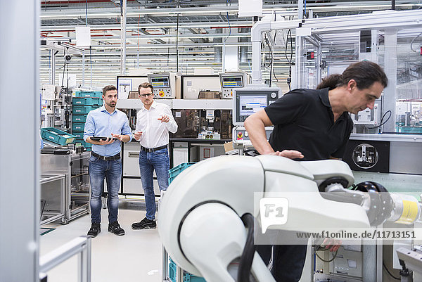 Man operating assembly robot in factory with two men in background supervising