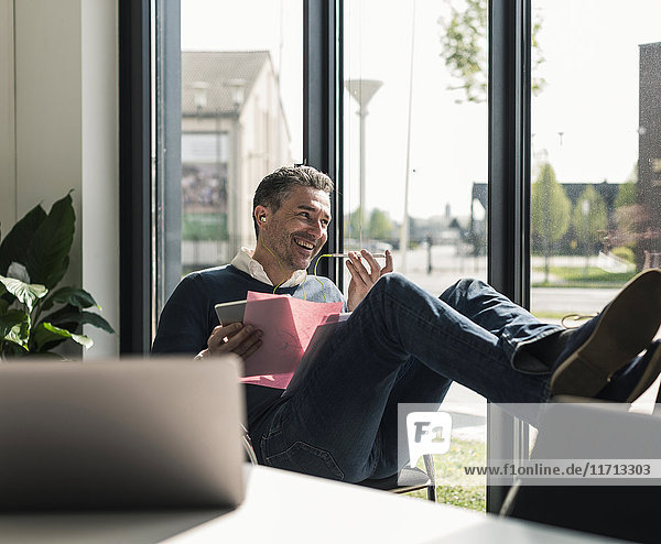 Lächelnder Geschäftsmann sitzt mit erhobenen Füßen in seinem Büro mit Smartphone und Kopfhörern.