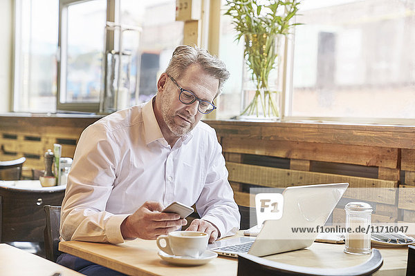 Mature businessman in cafe using laptop and cell phone