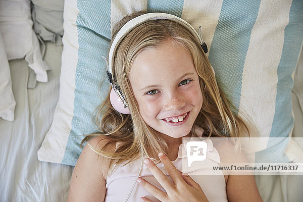 Porträt eines lächelnden kleinen Mädchens  das zu Hause auf dem Bett liegt und Musik mit Kopfhörern hört.