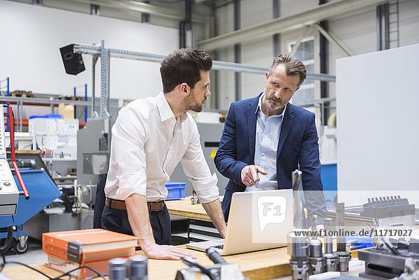 Two men at table in factory shop floor with laptop