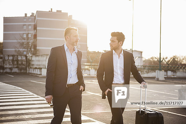 Two businessmen with rolling suitcase on parking place at sunset