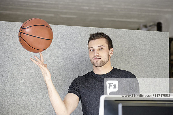 Junger Mann  der im Büro arbeitet und einen Basketball balanciert.
