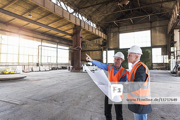 Two men with plan wearing safety vests talking in old industrial hall