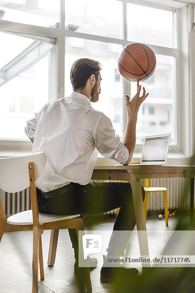 Rückansicht eines jungen Mannes am Schreibtisch mit Blick auf den Laptop beim Balancieren eines Basketballs am Finger