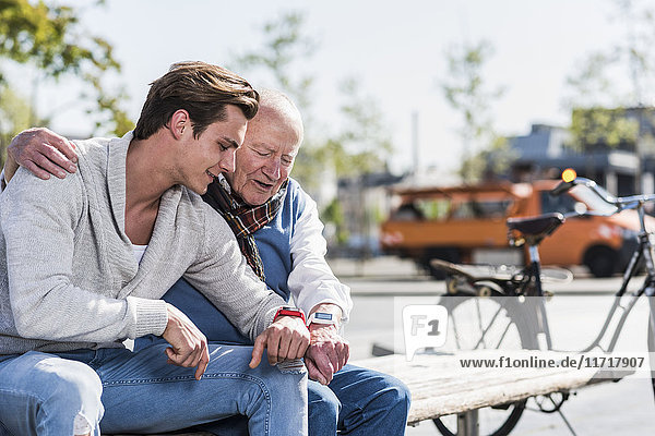 Senior Mann und erwachsener Enkel auf einer Bank mit Blick auf ihre Smartuhren