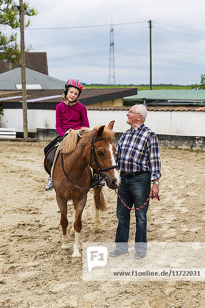 'Grandfather walking beside his granddaughter riding a horse in a stable  the teenager is wearing a pink helmet; North Rhein  Westphalia  Germany'