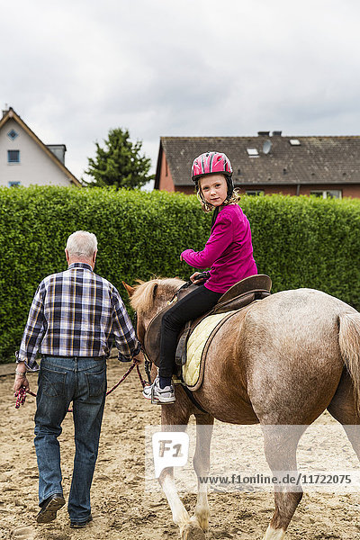 Großvater geht neben seiner Enkelin  die auf einem Pferd in einem Stall reitet  spazieren; der Teenager trägt einen rosa Helm; Nordrhein  Westfalen  Deutschland'.