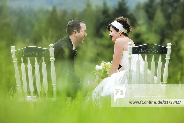 'A bride and groom sit side by side in chairs in a grass field  talking and laughing together; Oregon  United States of America'