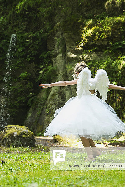 'A girl with blond hair wearing an angel costume with feather wings walks on lush grass in a park; Oregon  United States of America'