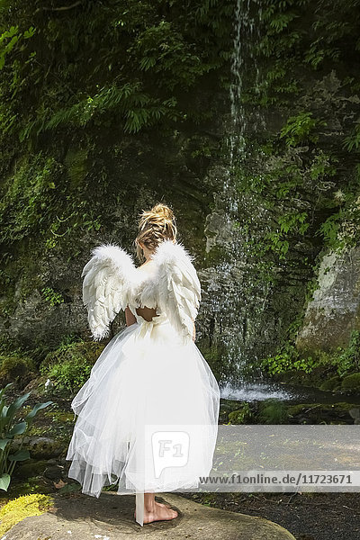 'A girl with blond hair wearing an angel costume with feather wings stands at the edge of a tranquil pool; Oregon  United States of America'