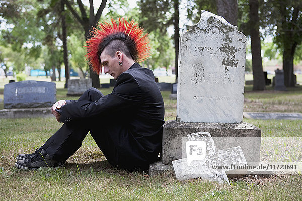 Ein junger Mann mit rotem Irokesenschnitt sitzt allein neben einem Grabstein auf einem Friedhof; Edmonton  Alberta  Kanada .