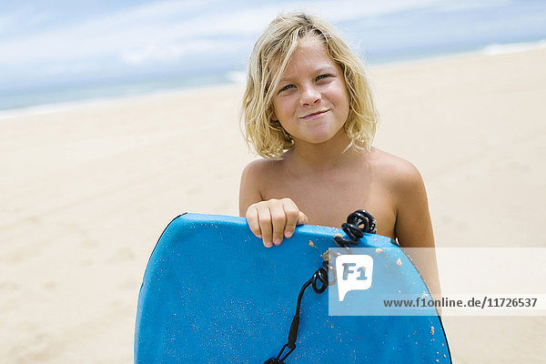 Junge (8-9) steht am Strand und hält ein Surfbrett